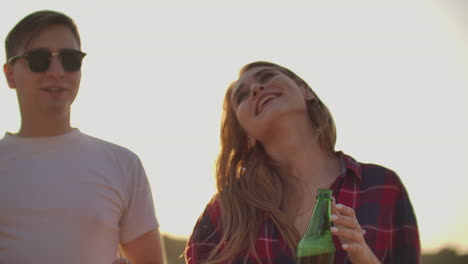 Loving-couple-is-enjoying-summer-open-air-party-with-friends.-Young-girl-is-dancing-with-beer-in-red-plaid-shirt-with-her-boyfriend-in-white-T-shirt-and-black-sun-glasses.
